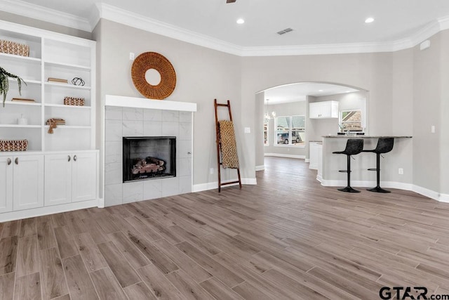 unfurnished living room featuring a tiled fireplace, crown molding, arched walkways, and light wood-type flooring