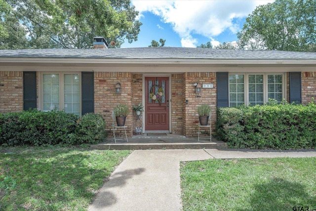 entrance to property featuring covered porch, a chimney, a shingled roof, a lawn, and brick siding