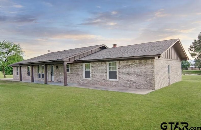 back of property at dusk with board and batten siding, a lawn, brick siding, and roof with shingles