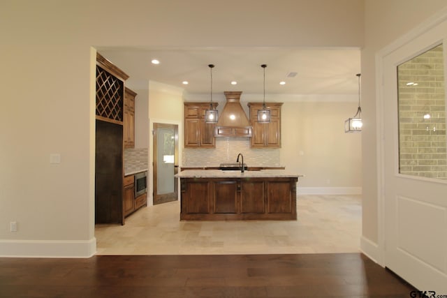 kitchen featuring tasteful backsplash, custom range hood, pendant lighting, light hardwood / wood-style floors, and a kitchen island with sink