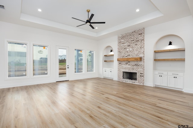 unfurnished living room featuring built in features, a fireplace, light hardwood / wood-style floors, a tray ceiling, and ceiling fan