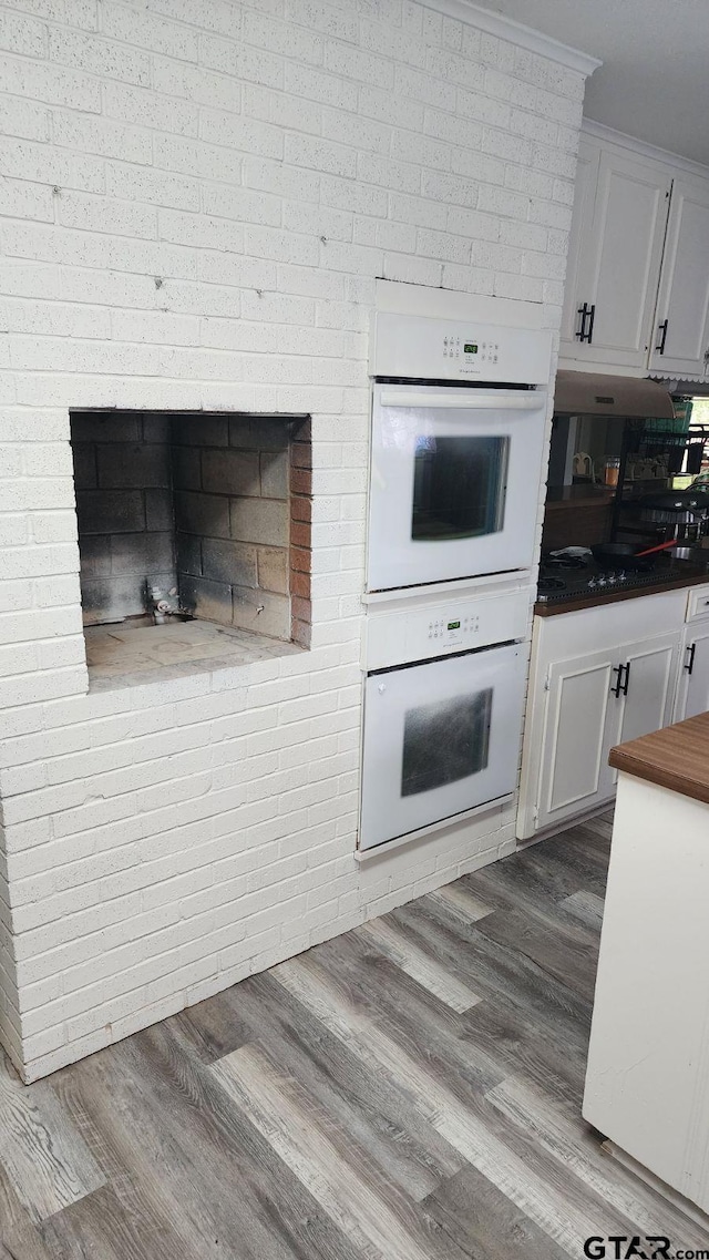 kitchen featuring a brick fireplace, wood-type flooring, white double oven, white cabinetry, and brick wall