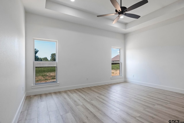 empty room with a raised ceiling, ceiling fan, plenty of natural light, and light hardwood / wood-style flooring