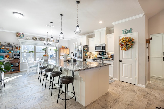 kitchen featuring appliances with stainless steel finishes, pendant lighting, white cabinets, dark stone counters, and a kitchen island with sink