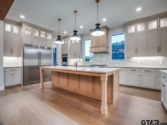 kitchen with stainless steel appliances, a center island with sink, decorative backsplash, light hardwood / wood-style flooring, and decorative light fixtures