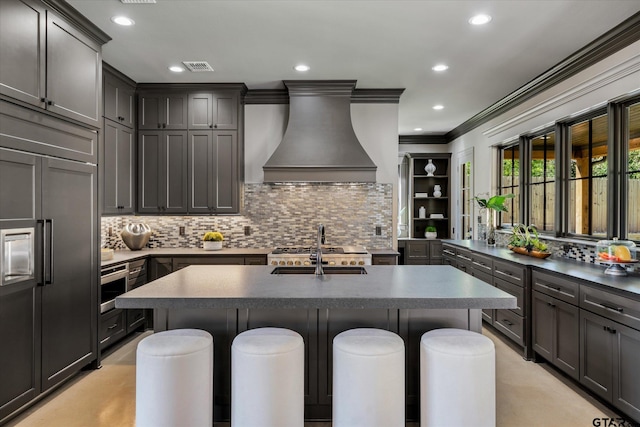 kitchen featuring a breakfast bar area, custom exhaust hood, paneled built in fridge, and an island with sink