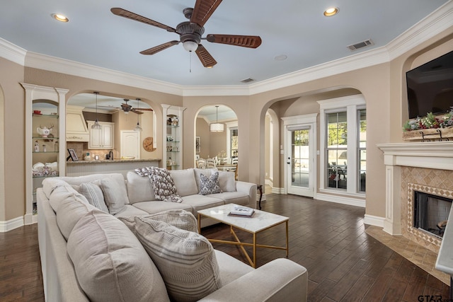 living room with a tiled fireplace, crown molding, dark wood-type flooring, and ceiling fan