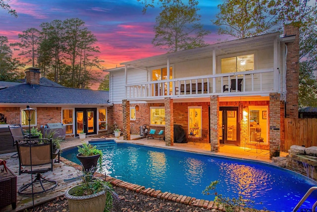 back house at dusk featuring a patio area, a balcony, a fenced in pool, and french doors