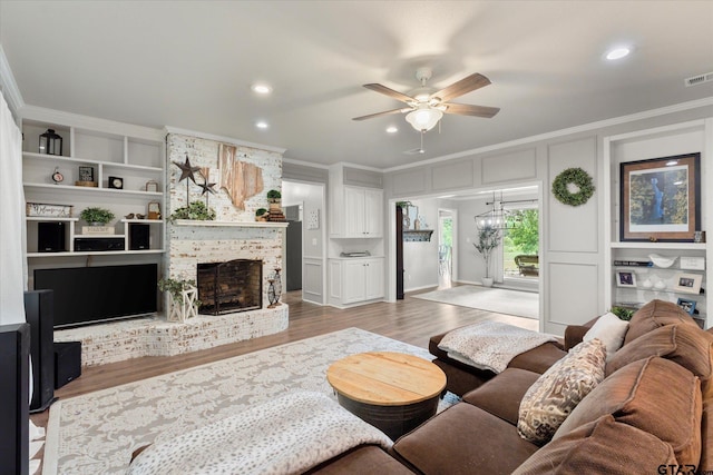 living room with ceiling fan, built in features, a brick fireplace, light hardwood / wood-style flooring, and crown molding