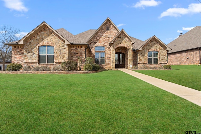 french country style house featuring a front lawn, brick siding, stone siding, and roof with shingles