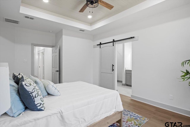 bedroom with ensuite bath, ceiling fan, a raised ceiling, a barn door, and wood-type flooring