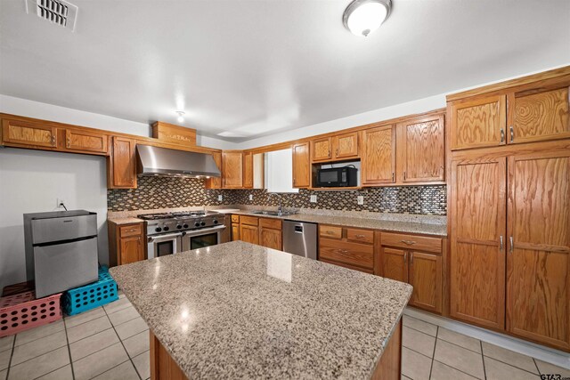 kitchen featuring light stone countertops, wall chimney range hood, light tile patterned flooring, a kitchen island, and appliances with stainless steel finishes