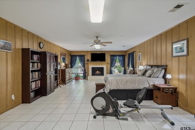 bedroom with wood walls, a warm lit fireplace, visible vents, a ceiling fan, and light tile patterned floors