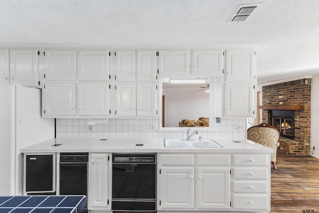 kitchen featuring tile countertops, white cabinetry, dishwasher, sink, and a brick fireplace