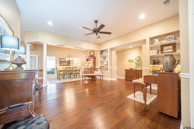 sitting room with built in shelves, hardwood / wood-style flooring, ceiling fan with notable chandelier, visible vents, and decorative columns