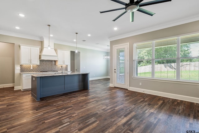 kitchen with dark wood-type flooring, a center island with sink, white cabinets, crown molding, and decorative light fixtures