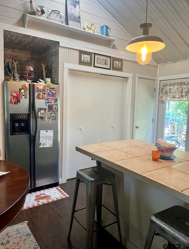 kitchen with wood walls, stainless steel refrigerator with ice dispenser, vaulted ceiling, tile counters, and a breakfast bar area