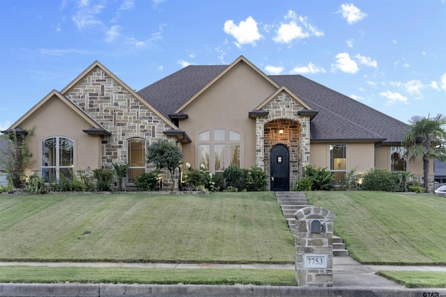 french country home featuring a shingled roof, stone siding, a front lawn, and stucco siding