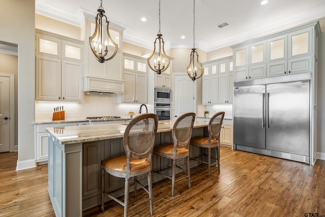 kitchen with an island with sink, built in appliances, light stone counters, and wood-type flooring