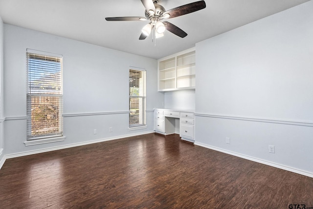 unfurnished office with built in shelves, ceiling fan, built in desk, and dark wood-type flooring