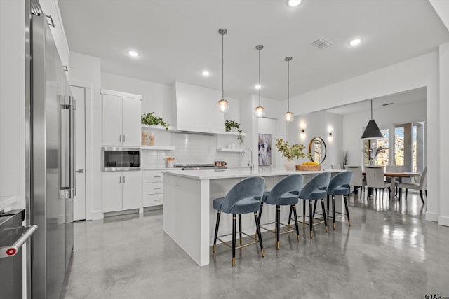 kitchen with white cabinetry, premium range hood, an island with sink, pendant lighting, and appliances with stainless steel finishes