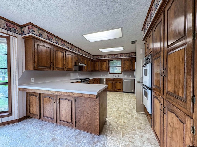 kitchen with a sink, a textured ceiling, appliances with stainless steel finishes, a peninsula, and under cabinet range hood