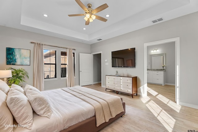 bedroom with connected bathroom, light hardwood / wood-style flooring, a raised ceiling, and ceiling fan