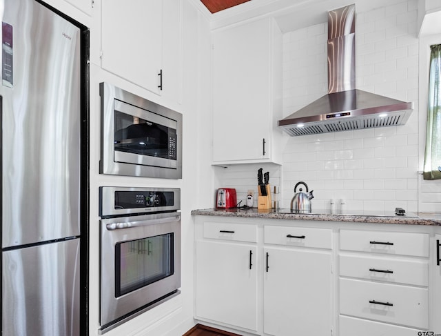 kitchen featuring light stone counters, tasteful backsplash, wall chimney range hood, white cabinetry, and appliances with stainless steel finishes