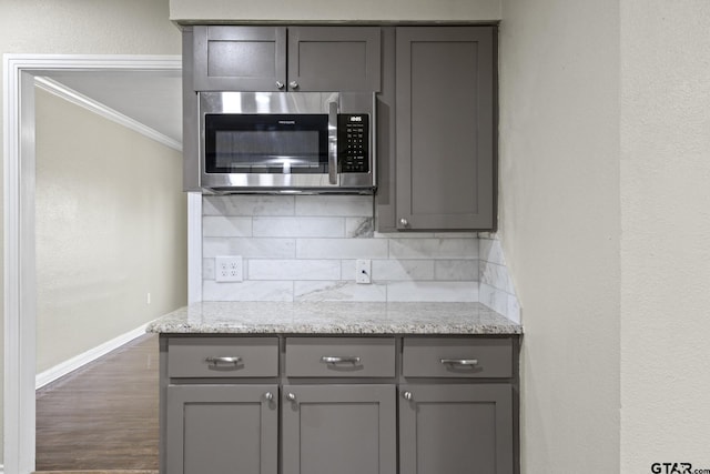 kitchen with dark wood-type flooring, gray cabinetry, light stone counters, crown molding, and backsplash