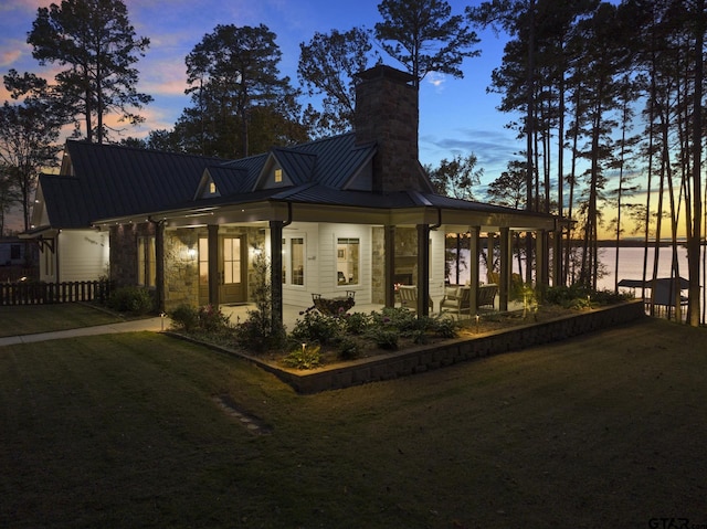 back of house at dusk featuring metal roof, a water view, fence, a lawn, and a standing seam roof