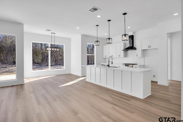 kitchen featuring hanging light fixtures, a kitchen island with sink, white cabinets, and wall chimney exhaust hood