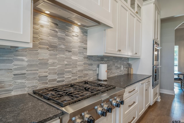 kitchen with white cabinetry, wall chimney exhaust hood, backsplash, dark stone counters, and hardwood / wood-style flooring