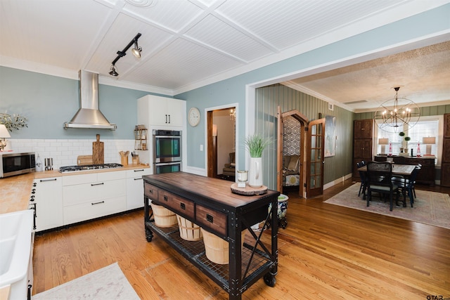 kitchen with stainless steel appliances, light countertops, white cabinets, wall chimney range hood, and light wood-type flooring