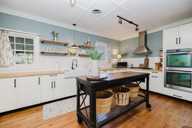 kitchen with light wood finished floors, a sink, stainless steel appliances, wall chimney range hood, and butcher block counters