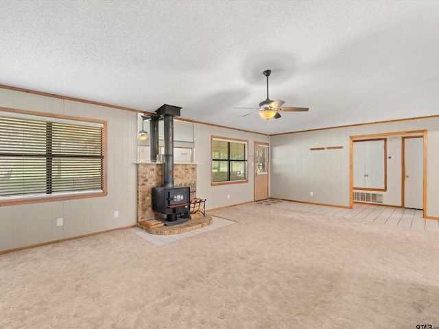 unfurnished living room featuring a wood stove, a textured ceiling, light carpet, ceiling fan, and crown molding