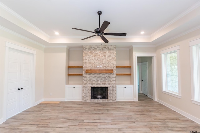 unfurnished living room featuring a raised ceiling, ornamental molding, light wood-type flooring, a brick fireplace, and ceiling fan