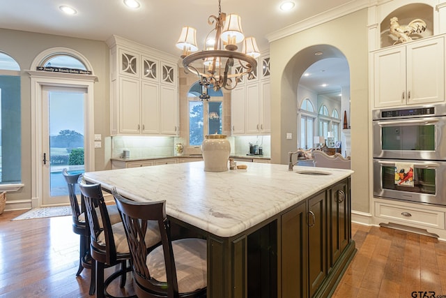 kitchen with sink, an island with sink, stainless steel double oven, dark hardwood / wood-style floors, and pendant lighting