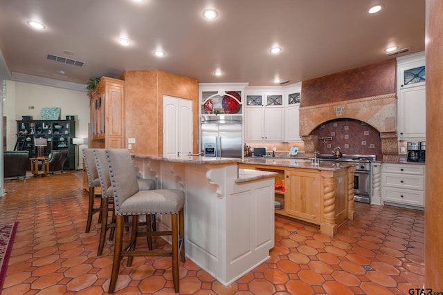 kitchen featuring high end appliances, a kitchen island with sink, decorative backsplash, light stone counters, and a breakfast bar area