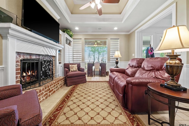 living room featuring a brick fireplace, light tile patterned floors, ornamental molding, ceiling fan, and a raised ceiling