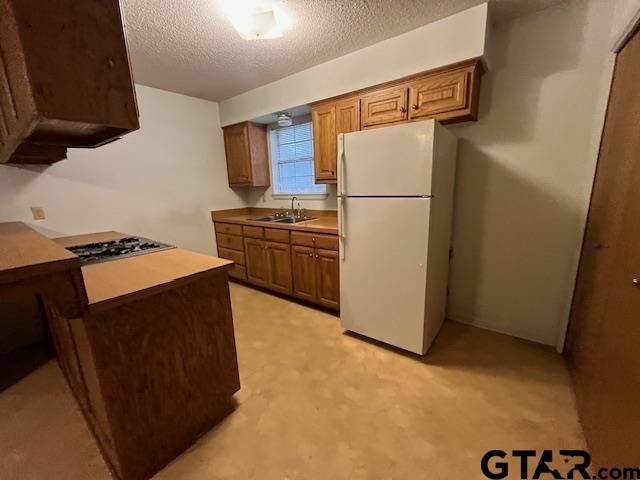 kitchen with a sink, a textured ceiling, freestanding refrigerator, stainless steel gas stovetop, and brown cabinetry