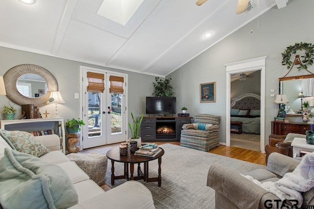 living room with a warm lit fireplace, vaulted ceiling with skylight, a ceiling fan, wood finished floors, and french doors