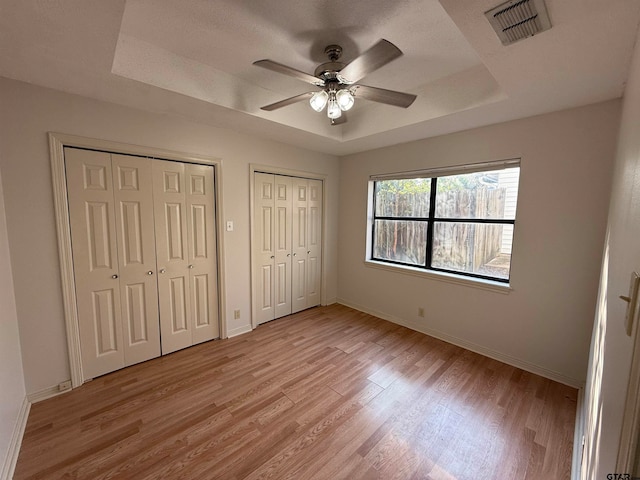 unfurnished bedroom featuring multiple closets, light wood-type flooring, a tray ceiling, and ceiling fan