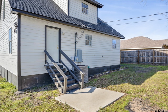 rear view of property featuring entry steps, a shingled roof, central AC unit, a lawn, and fence