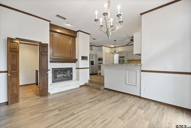 kitchen with a stone fireplace, light hardwood / wood-style floors, appliances with stainless steel finishes, ceiling fan with notable chandelier, and ornamental molding