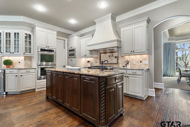 kitchen with arched walkways, stainless steel double oven, white cabinetry, custom range hood, and crown molding