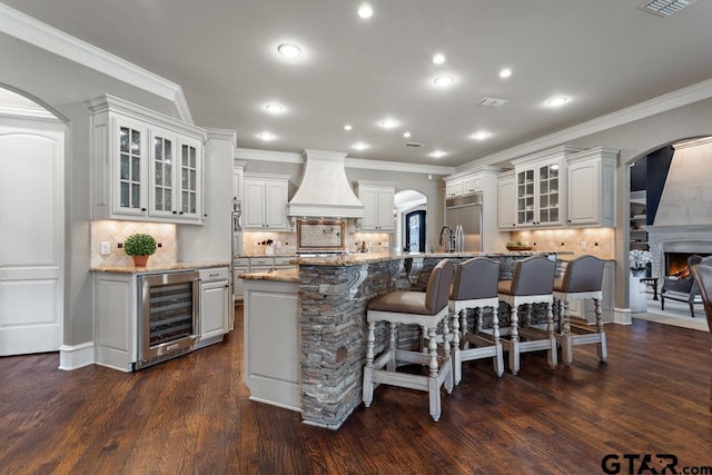 kitchen featuring wine cooler, arched walkways, custom exhaust hood, stainless steel built in fridge, and a kitchen bar