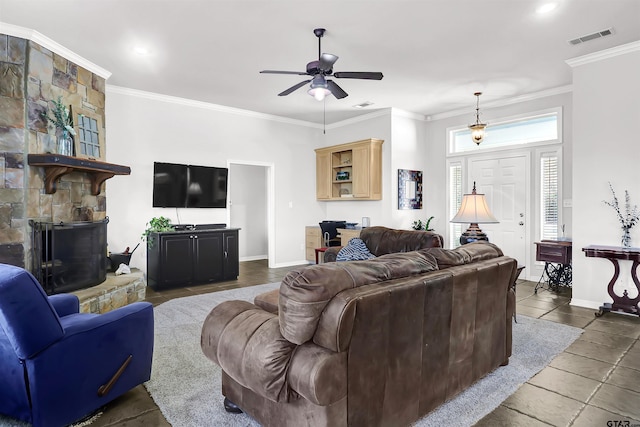 living room featuring dark tile patterned flooring, ceiling fan, crown molding, and a stone fireplace