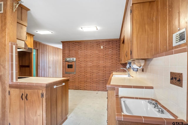 kitchen with tile counters, kitchen peninsula, sink, brick wall, and stainless steel oven