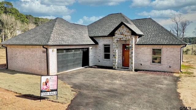 french country inspired facade with aphalt driveway, an attached garage, brick siding, and roof with shingles