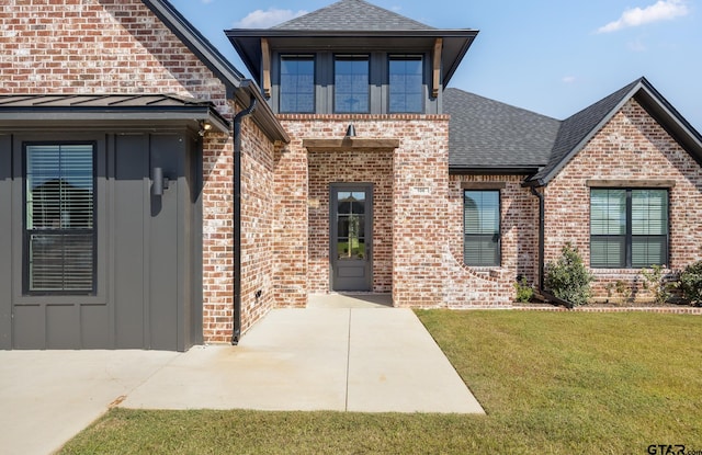 view of exterior entry featuring a yard, brick siding, and roof with shingles